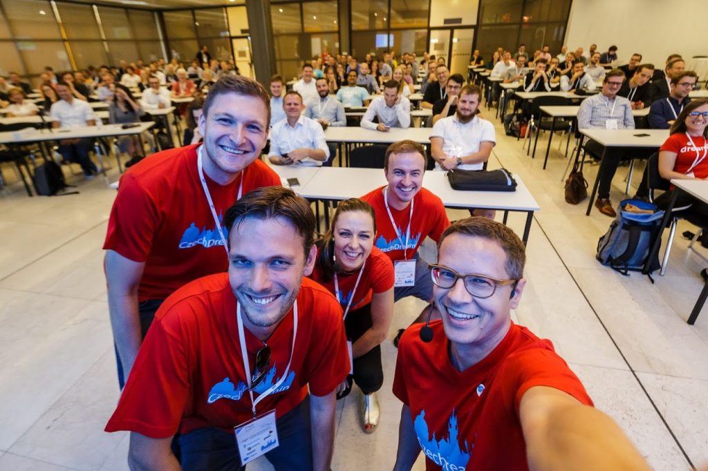 The Czech Dreamin organising crew smiling massively with attendees sitting at tables behind them, filling the room