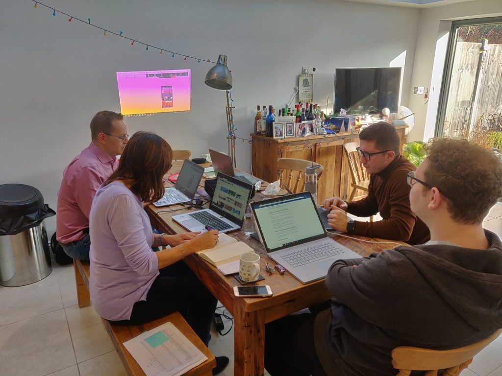 4 people crowded around a table, looking at laptops, at someone's home