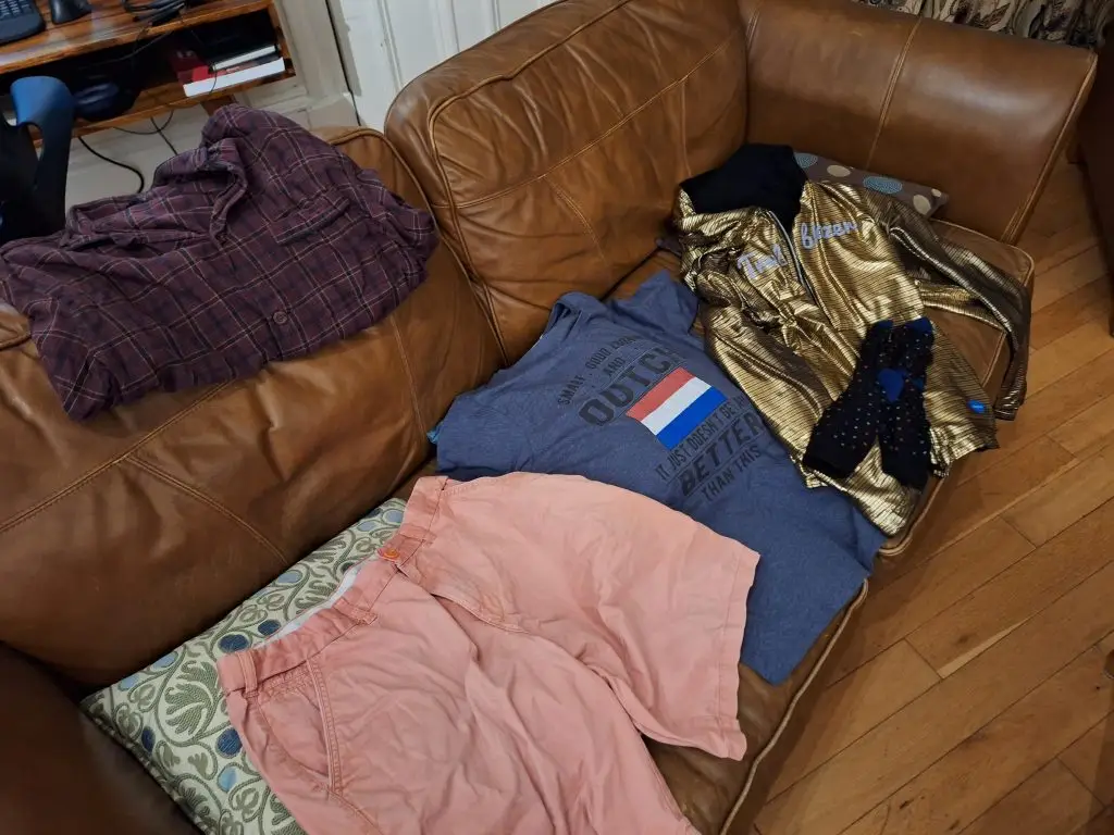 Picture of a chequered pyjama top, pink shorts, a T-Shirt with the Dutch flag, and my golden hoodie, all lying on a sofa 